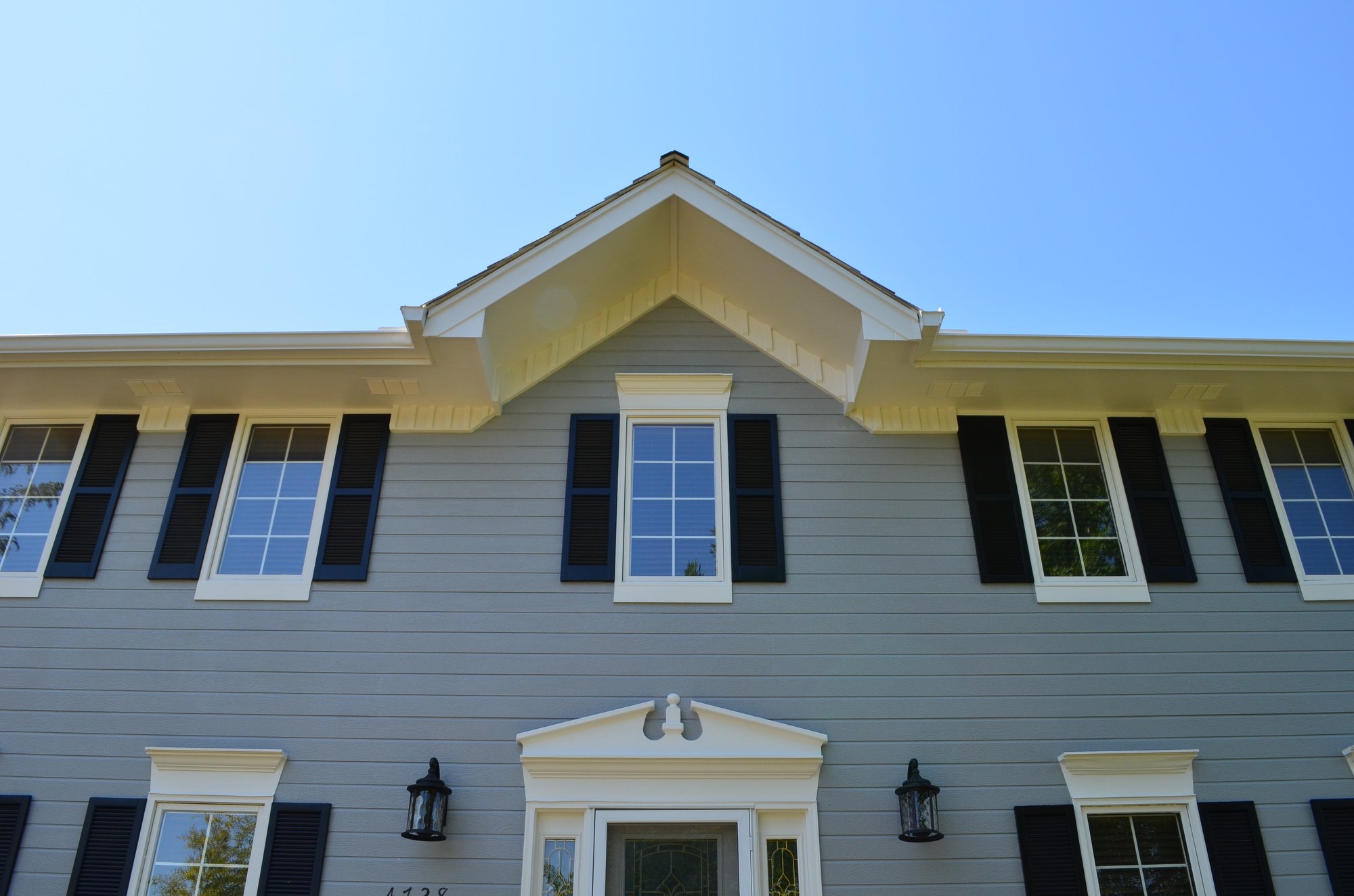 View looking up at the front of a light blue home with darker shutters, in front of the bright blue sky.