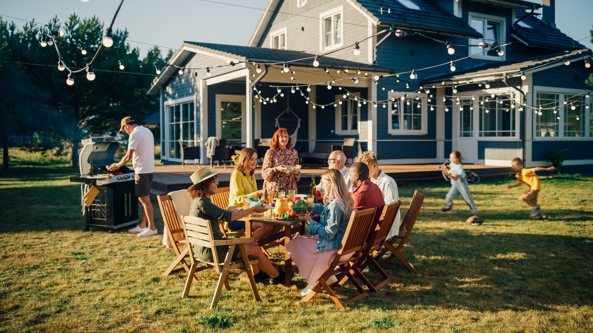 Family gathered around a table in a backyard with lights strung above them and blue exterior of home behind.